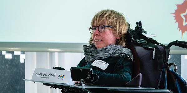 Eine Frau im Rollstuhl steht vor einer Präsentation. Vor ihr ist ein Schild mit ihrem Namen 