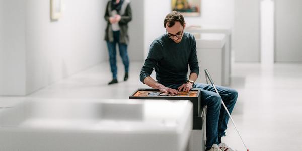 Blinder Mann sitzt im Ausstellungsraum und tastet mit einen Kunstgegenstand ab.