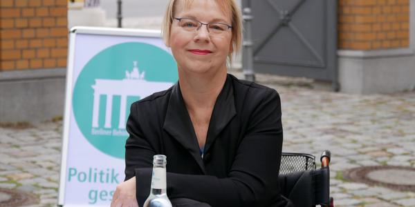 Eine Frau im Rollstuhl sitzt an einem Tisch. Sie hat kurze blonde Haare und trägt eine schwarze Bluse.
