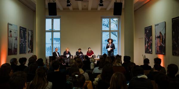 In einem dunkeln Raum sitzen 4 Personen auf einem Podium. Rechts daneben steht eine Moderatorin mit einem Mikrofon.