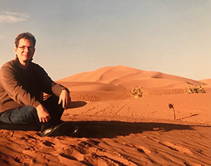 Foto von Michael Spörke. Er sitzt in der Sahara im Wüstensand bei Sonnenuntergang.