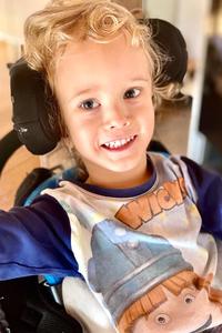 Der kleine Korbinian, ein blond gelockter Junge, sitzt in einem Therapiestuhl und lacht.
