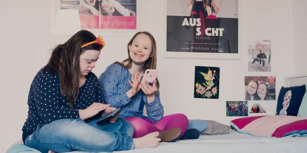 Zwei Mädchen sitzen auf einer Liege, an der Wand sind Poster angebracht