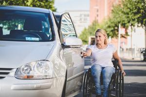 Eine Frau im Rollstuhl steht neben einem Auto und öffnet die Tür an der Fahrerseite.