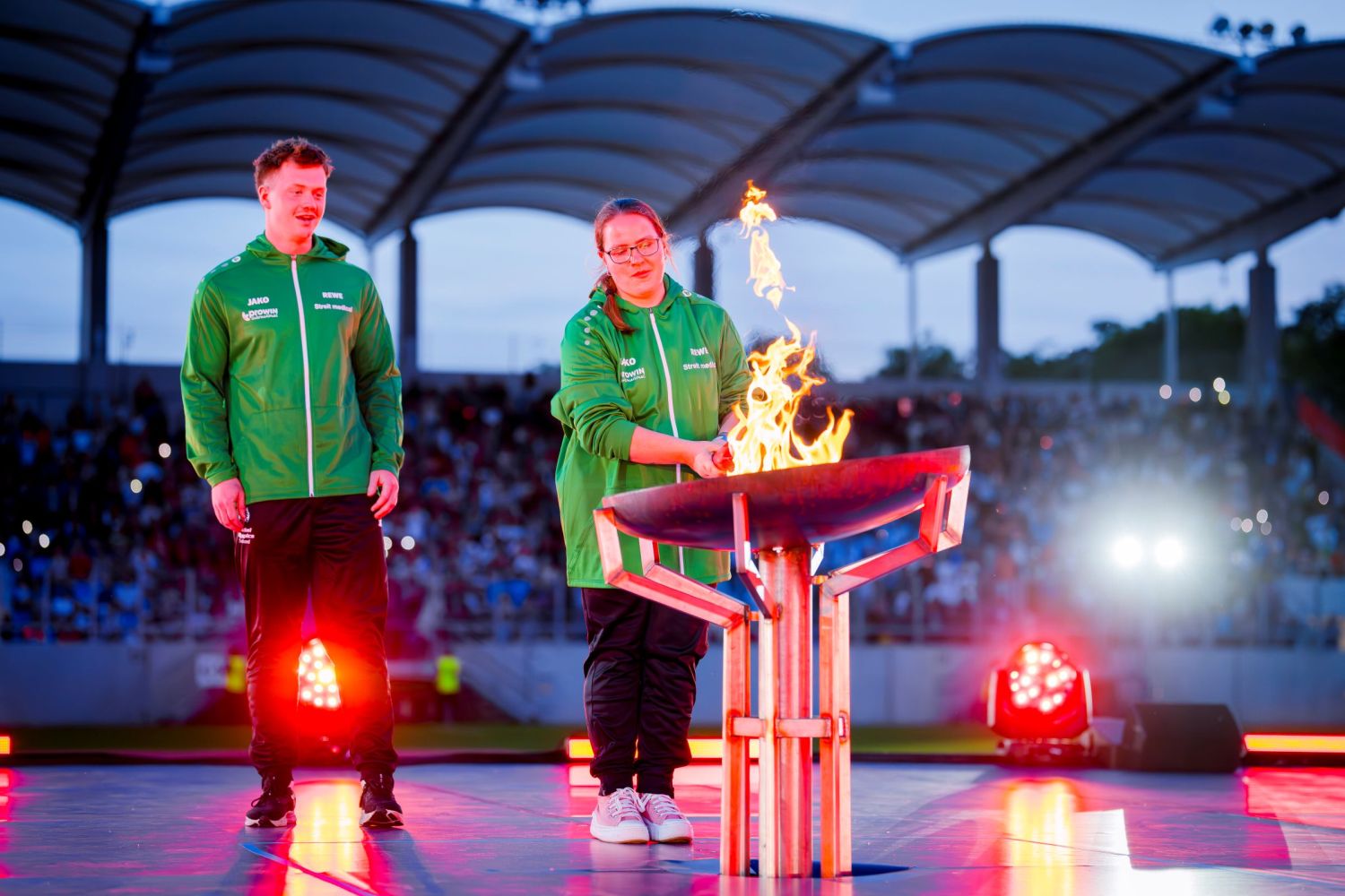 Ein junger Mann und eine junge Frau mit grüner Sportkleidung entzünden das Feuer an einer Feuerschale