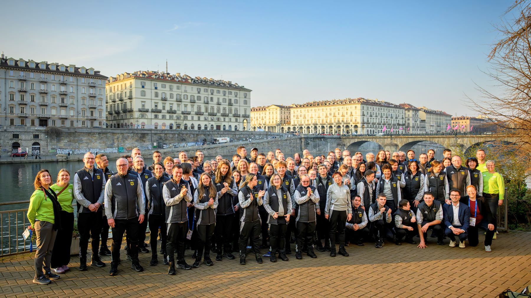 Große Gruppe von Sportlerinnen und Sportlern vor der Silhouette mehrgeschossiger gelblicher Häuser