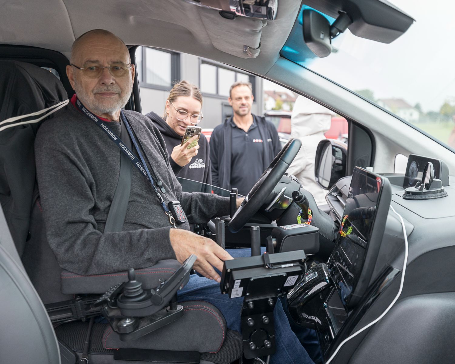 Ein Mann sitzt am Lenkrad eines Autos, das mit weiterer Technik zum Fahren durch mobilitäseingeschränkte Fahrer umgebaut wurde. Dabei schauen weitere Veranstaltungsteilnehmer zu.