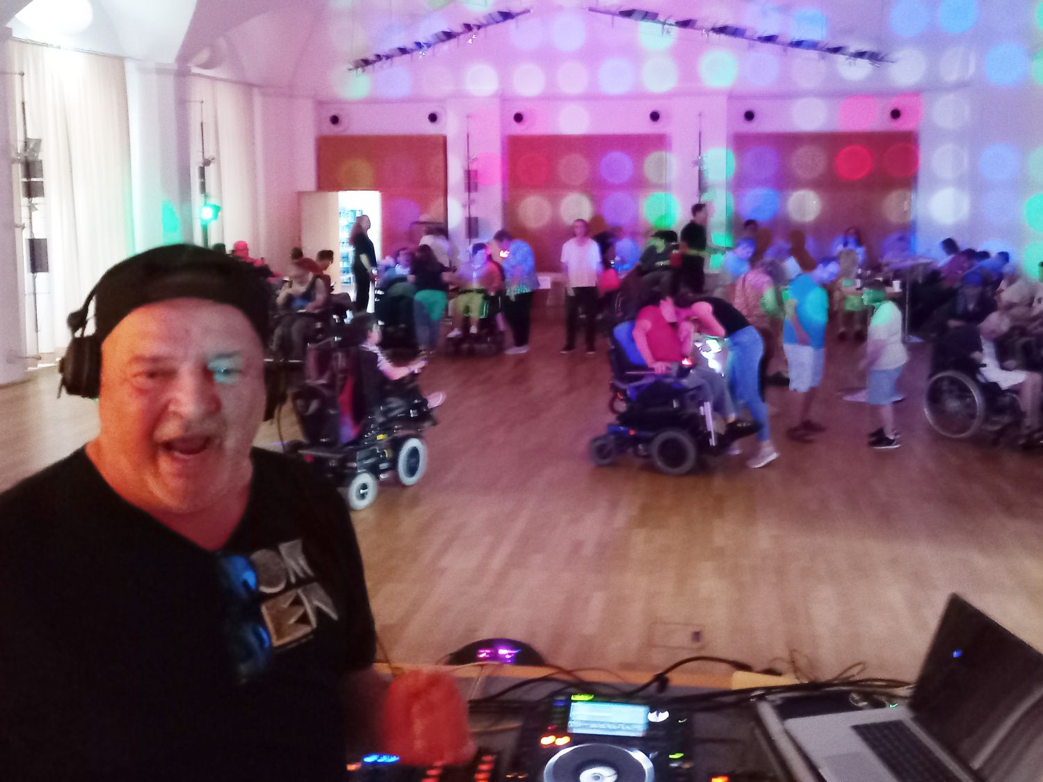 Links ein Mann mit Kopfhörern, Blick in den Raum mit tanzendenden Paaren, auch mit Rollstuhl bei Partylicht