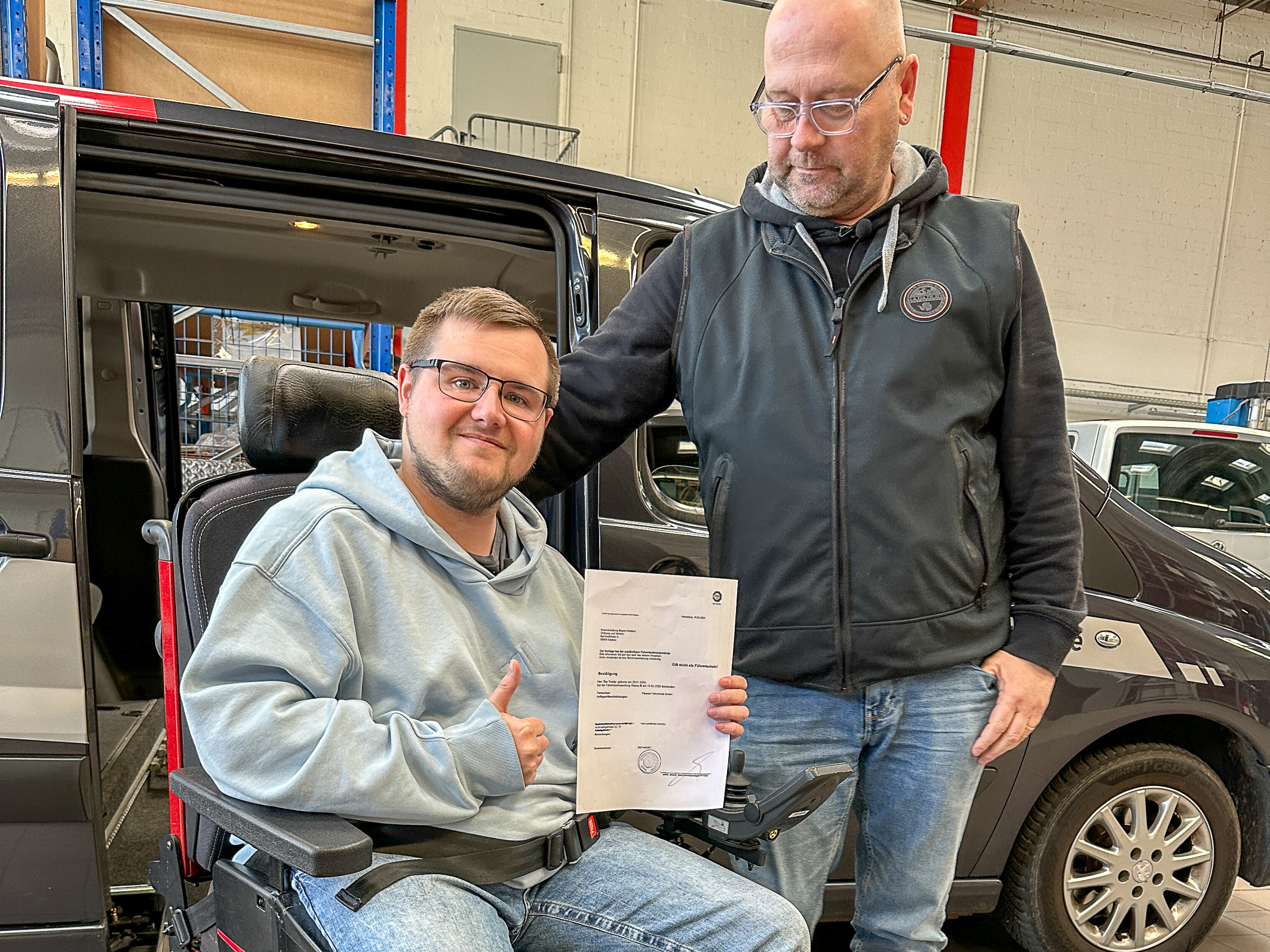 Junger Mann mit Brille und grauer Kleidung sitzt auf einem nach außen gedrehten Autositz und zeigt eine Urkunde. Neben ihm steht sein Fahrlehrer, ein älterer Mann.