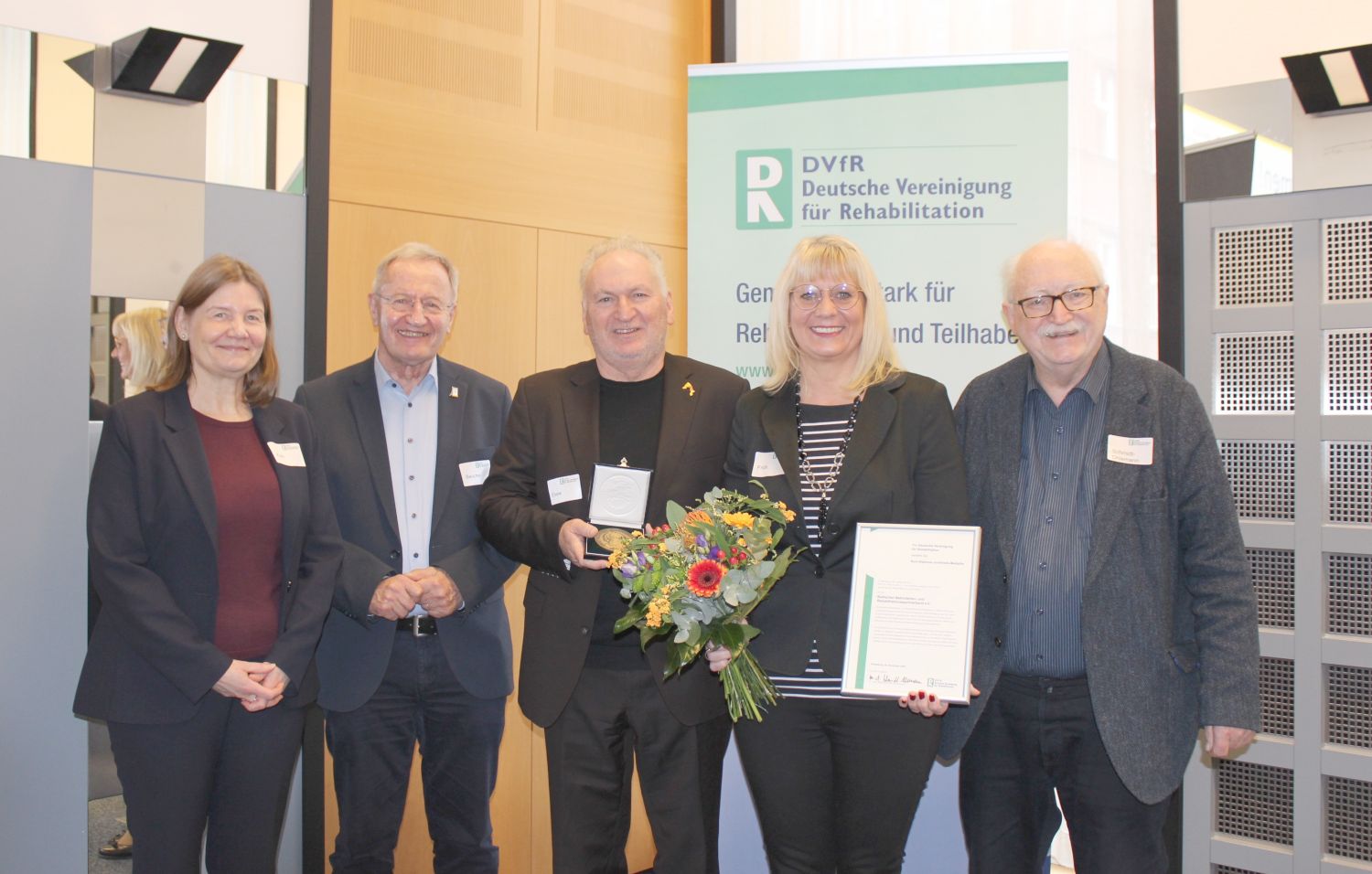 Drei Männer und eine frau stehen vor einer großen Tafel mit Vereinssymbol. Die frau hält einen Blumenstrauß und eine Urkunde in der Hand