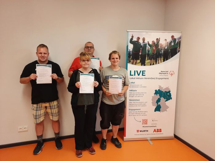 Vier Personen mit ihren Urkunden vor dem Schild der Schulungsreihe