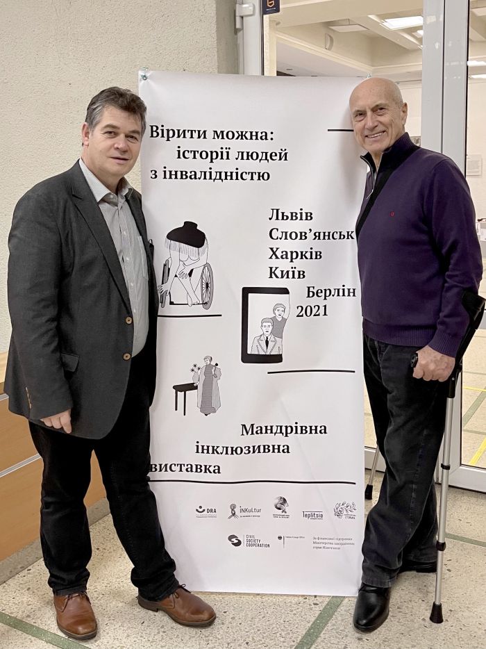 Zwei Männer und dazwischen ein Aufsteller am Eingang zur Ausstellung