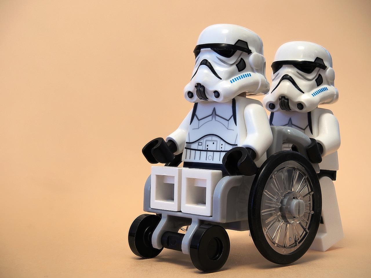 Ein Rollstuhlfahrer wird geschoben, beide im Look von Star Wars Truppen (Legofiguren).
