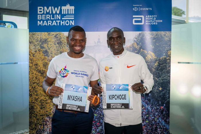 Zwei dunkelhäutige Sportler vor einer Tafel mit der Ankündigung de sMarathon