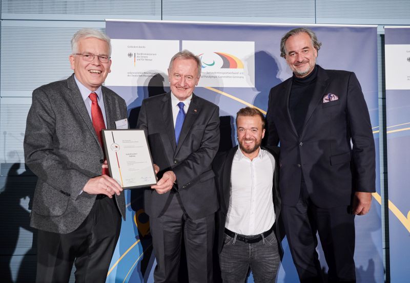 Drei Männer vor einer Tafel. Zwei Männer halten die Ehrenurkunde