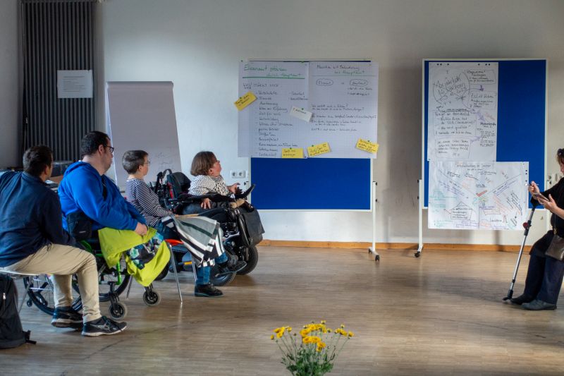 Gruppe vonn Personen, auch mit Rollstuhl links im Bild. Rechts zwei Tafeln mit Notizen.