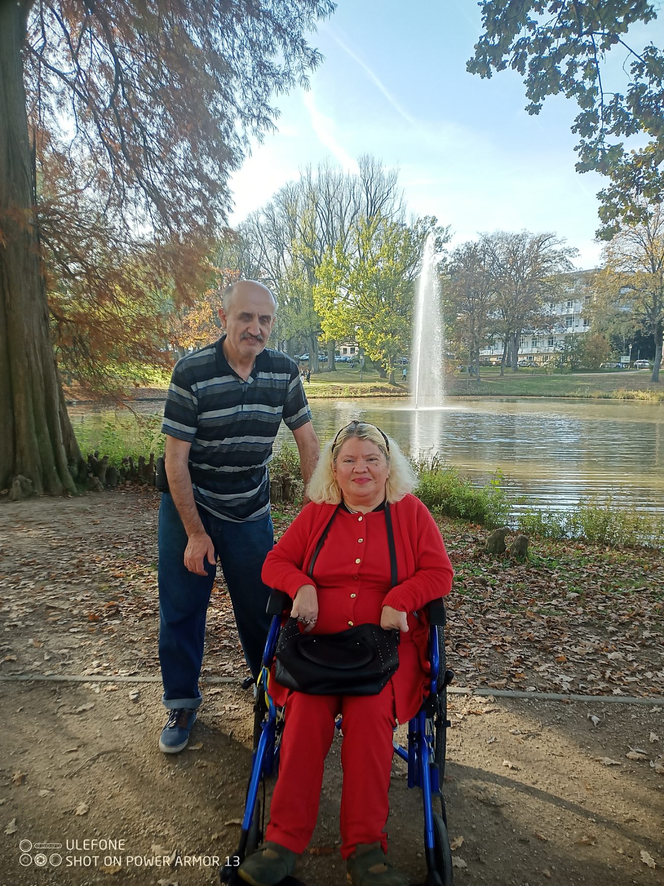 Frau mit Rollstuhl, dahinter steht ein Mann. Im Hintergrund ist ein Springbrunnen zu sehen