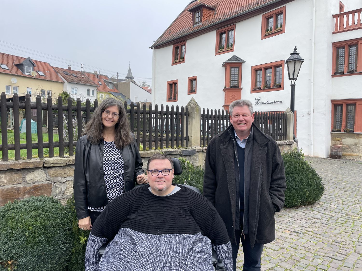 Person im Rolsltuhl mit einer Frau und einem  Mann vor dem Heimatmuseum