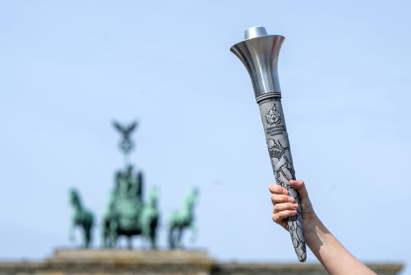 Eine Fackelaus Metall wird von einer Hand in de Höhe gehalten. Im Hintergrund die  Quadriga des Brandenburger Tors