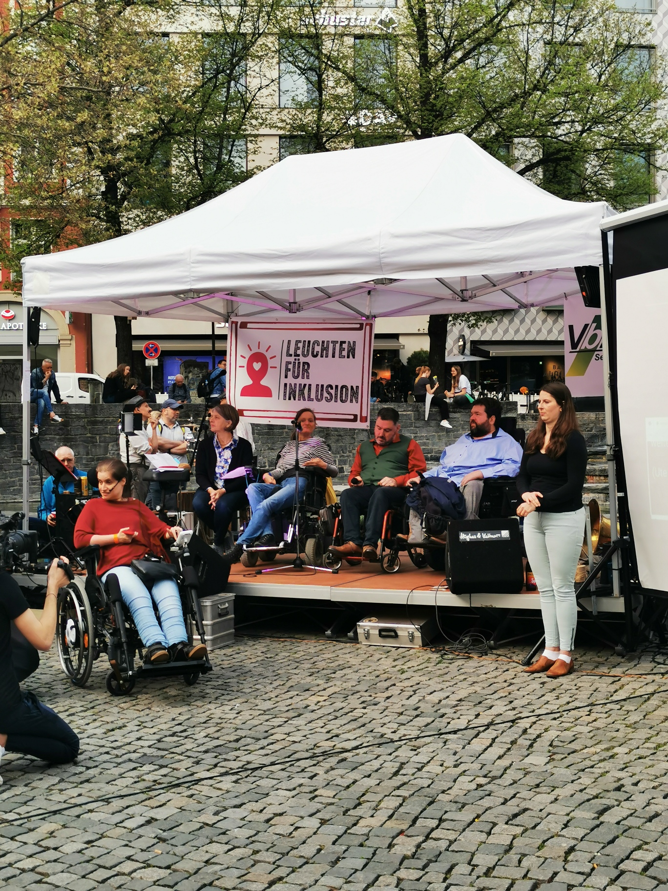 Menschen mit und ohne Rollstuhl unter einem offenen Zelt, die Plakate zeigen.