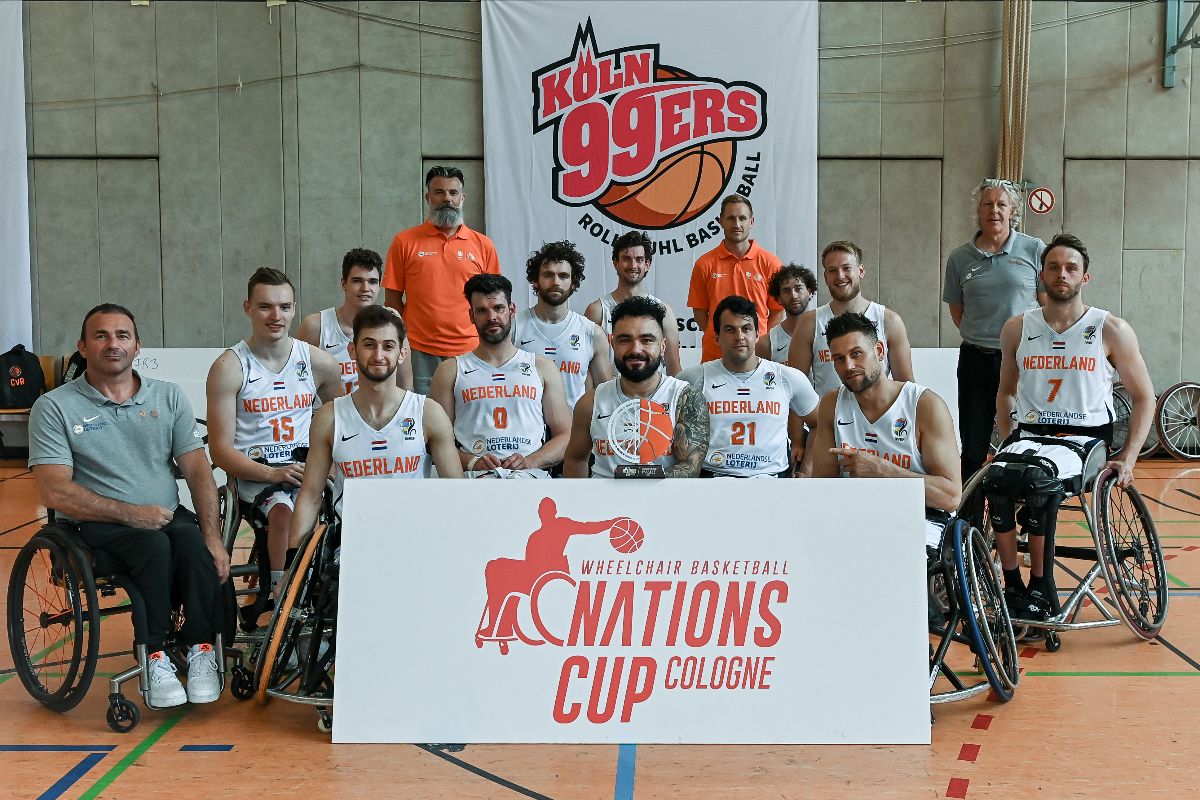 Rollstuhlbasketballer in zwei Reihe vor Wand mit dem Köln 99er Logo an der Rückand der Halle