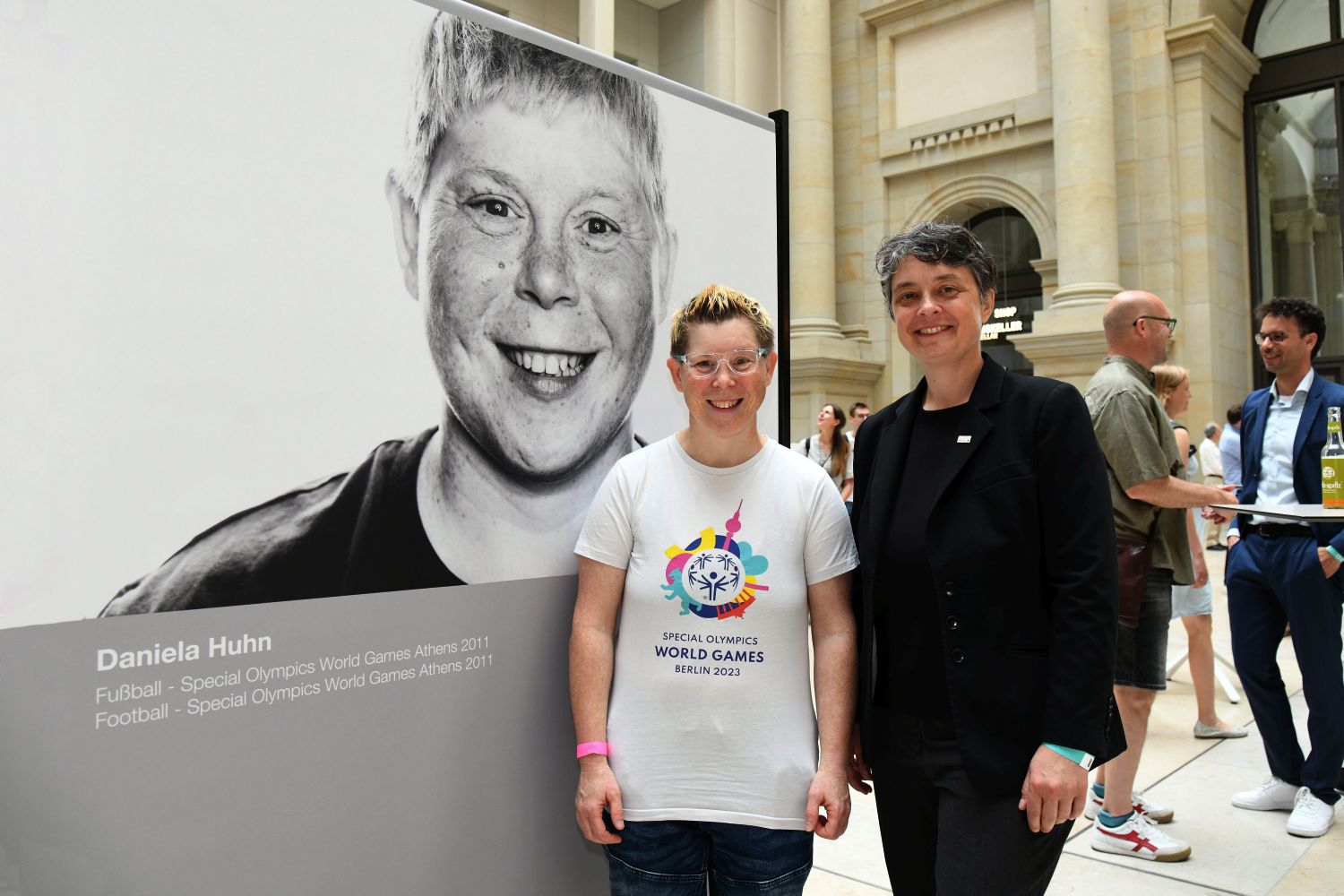 Daniela Huhn  mit Stattssekretärin vor einem Portraitfoto der Athletin