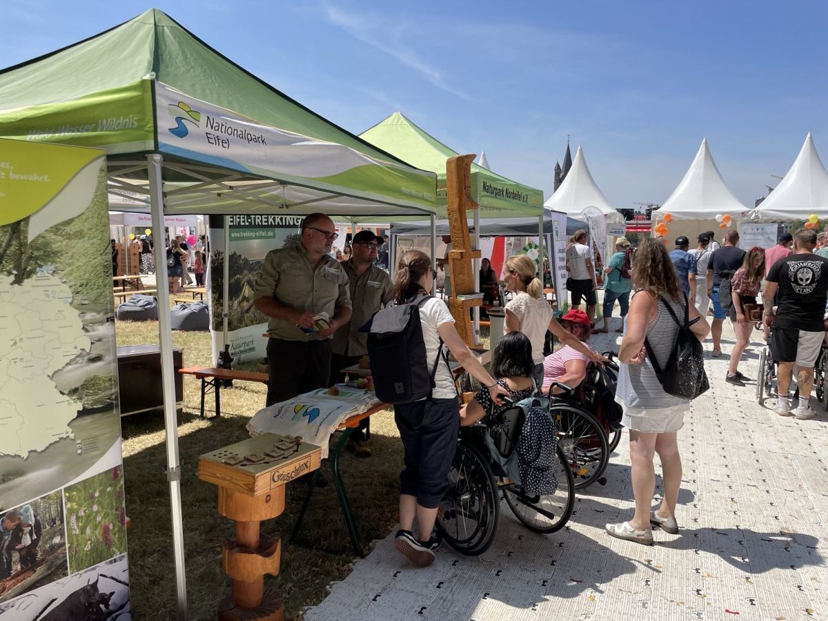 Stand mit Broschüren unter einem grünen Zeltdach mit Menschen davor