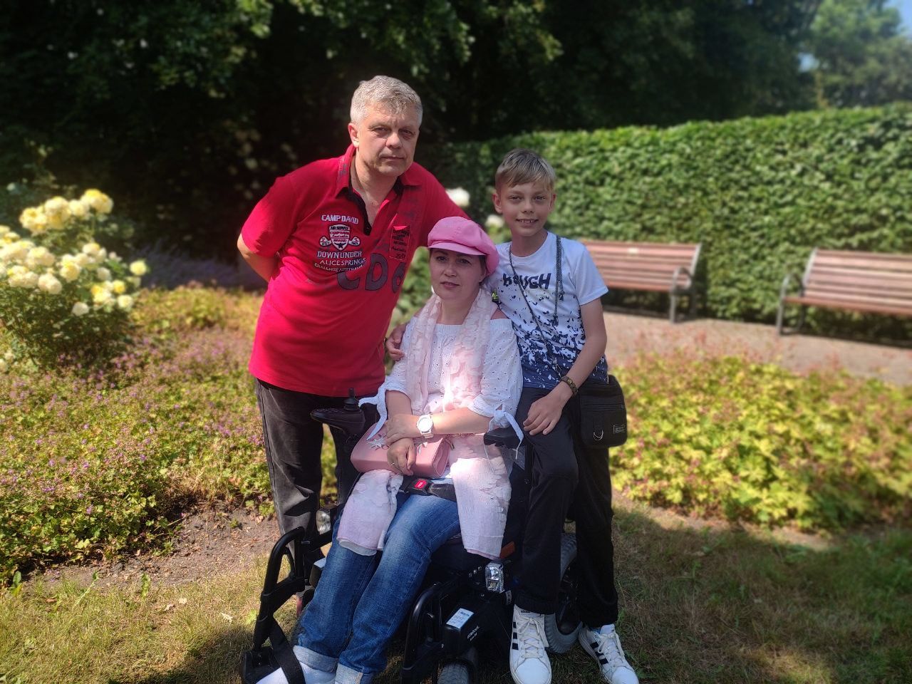 Junge Frau im Rollstuhl sitzend, links daneben stehend ein Mann, rechts daneben ein 13jähriger Junge