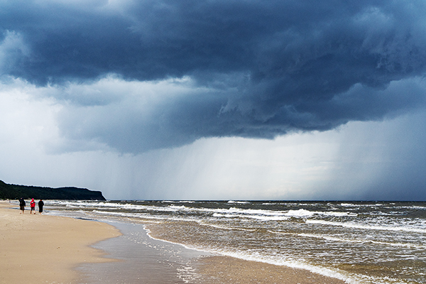 Dunkle Wolken über dem Meer, drei Menschen in der Ferne gehen den Strand entlang
