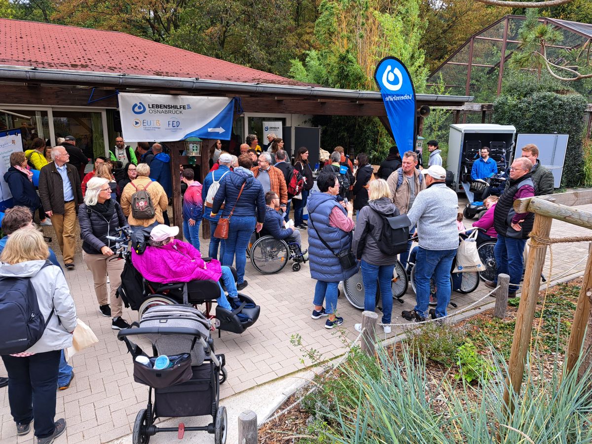 Viele Personen,  auch mit Rollstuhl, auf einem Platz vor einem eingeschossigen Gebäude