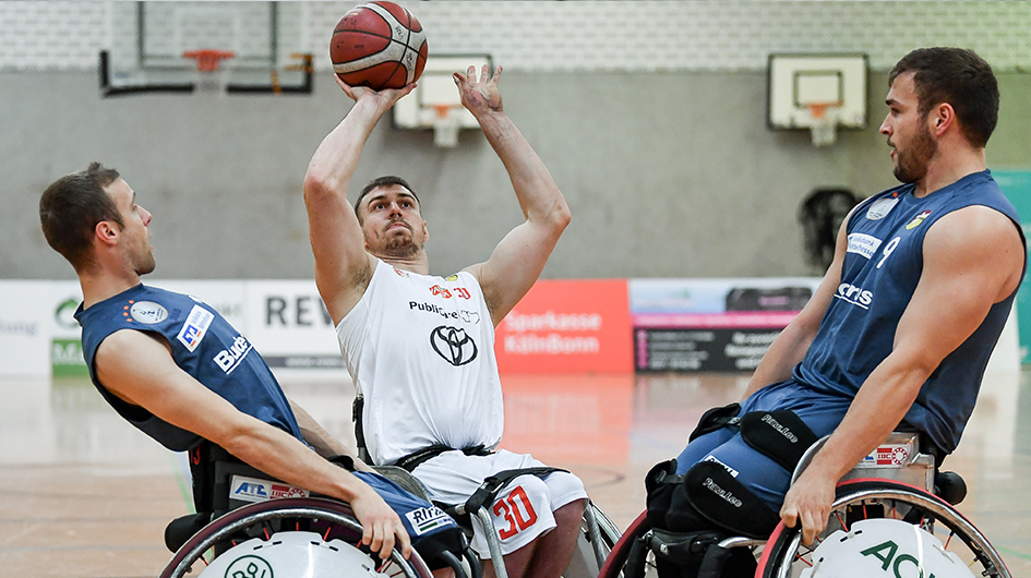 Drei Rollstuhlbasketballer von zwei Mannschaften im Kampf um den Ball. Ein Spieler um einen Wurf in den Korb bemüht