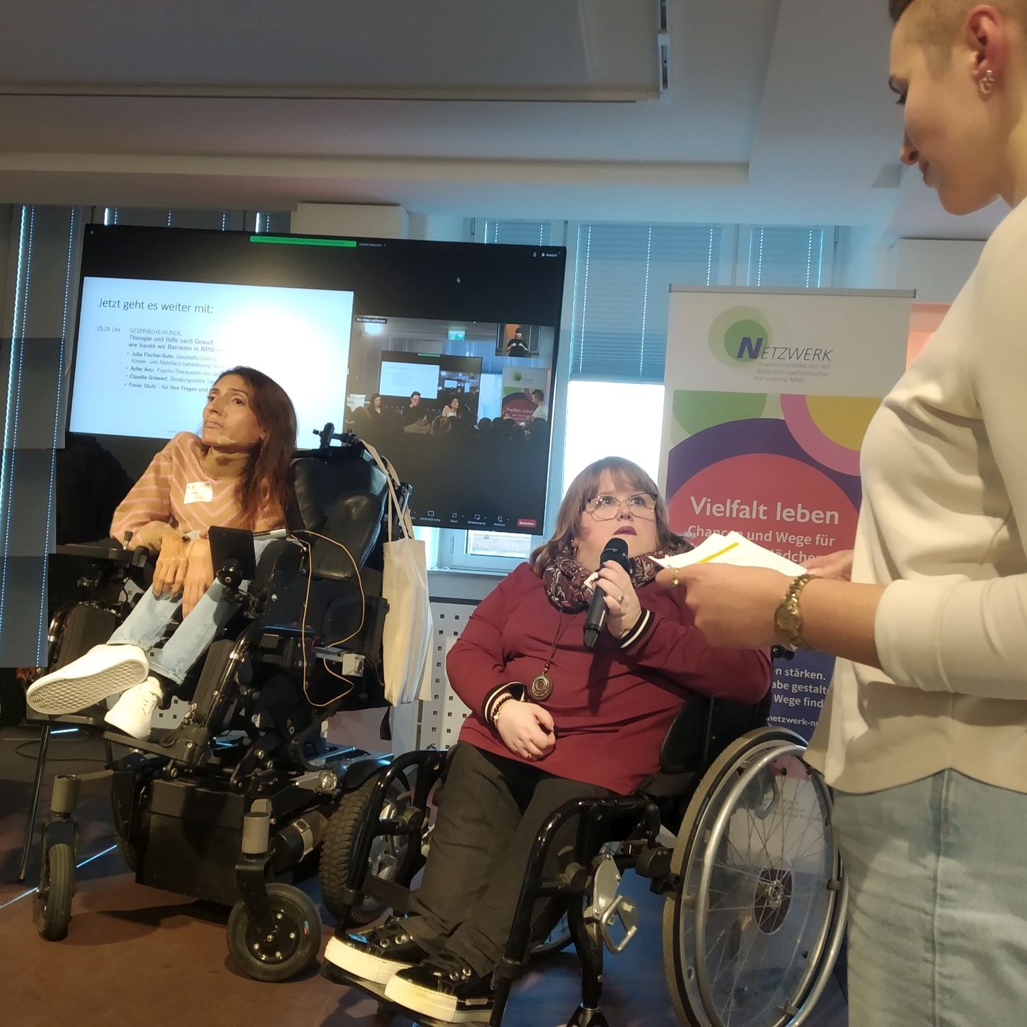 Auf dem Podium zwei Frauen eine hält Mikrofon nd spricht zur Moderatorin. Im Hintergrund Präsentation und Netzwerklogo