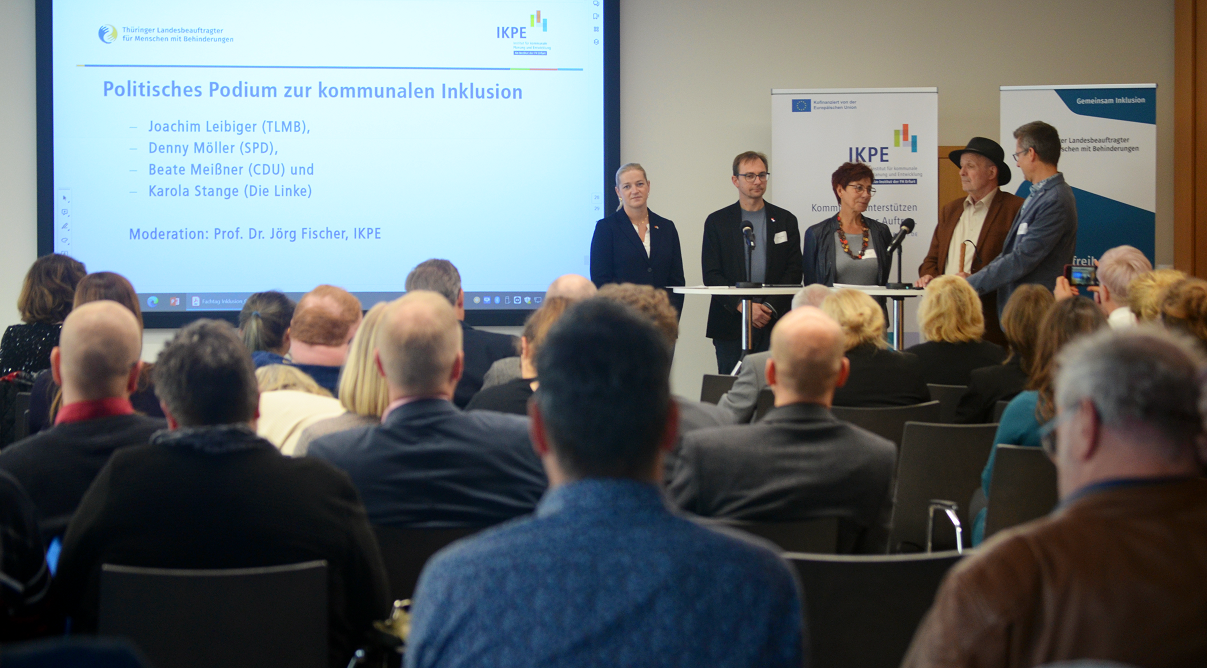 Teilnehmerinnen un d Teilnehmer im Raum blicken zu  einer Personengruppe auf einer Bühne.