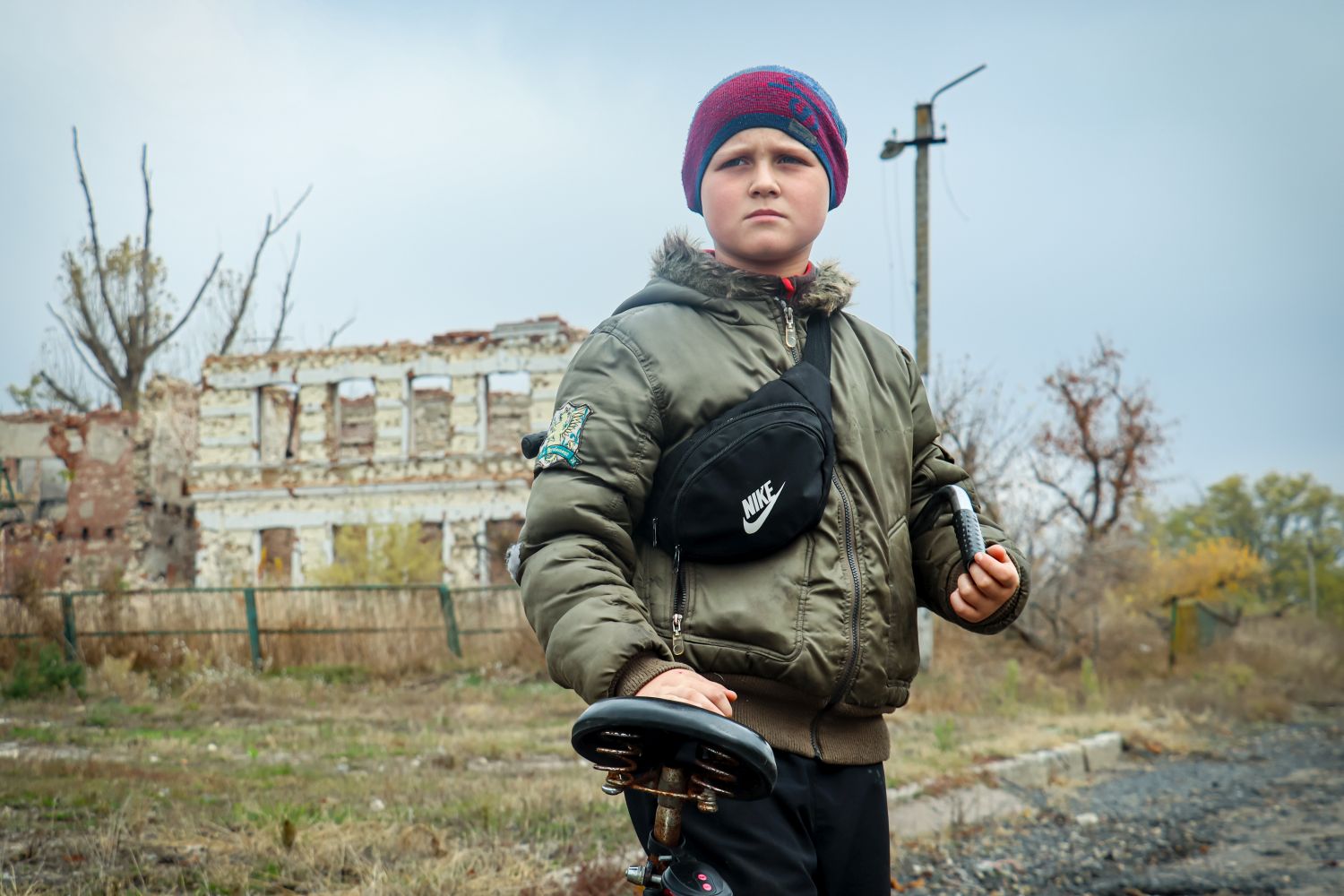 Ein elfjähriger Junge in dunkler Kleidung mit einem Minensuchgerät vor einem zerstörtem Wohnhaus