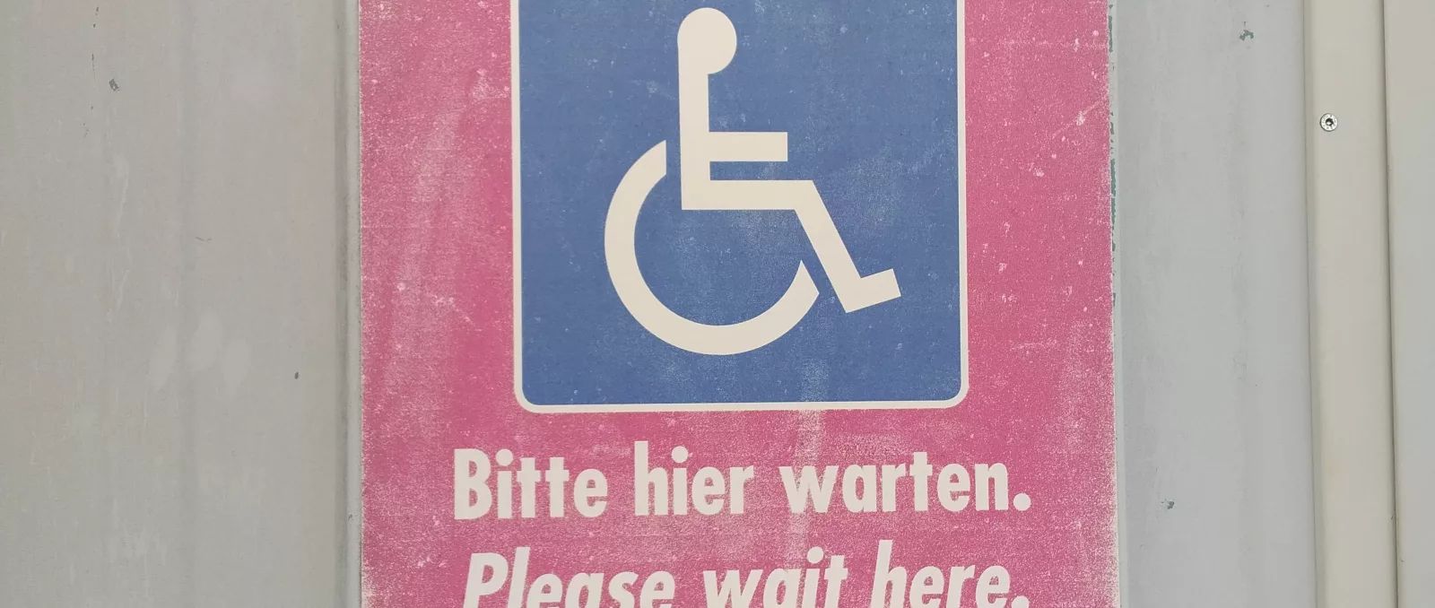 Schild mit Rollizeichen: Bitte hier warten. Please wait here.