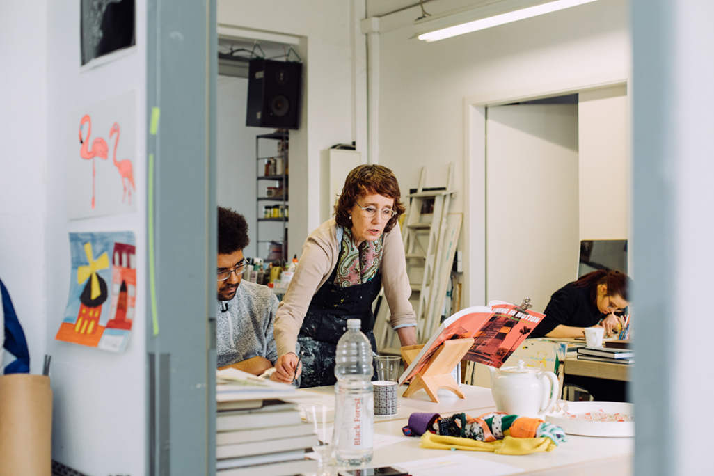 Zwei Personen sitzenam Tisch,  beugen ihre  Köpfe und arbeiten, eine Frau schaut ihnen über die Schulter