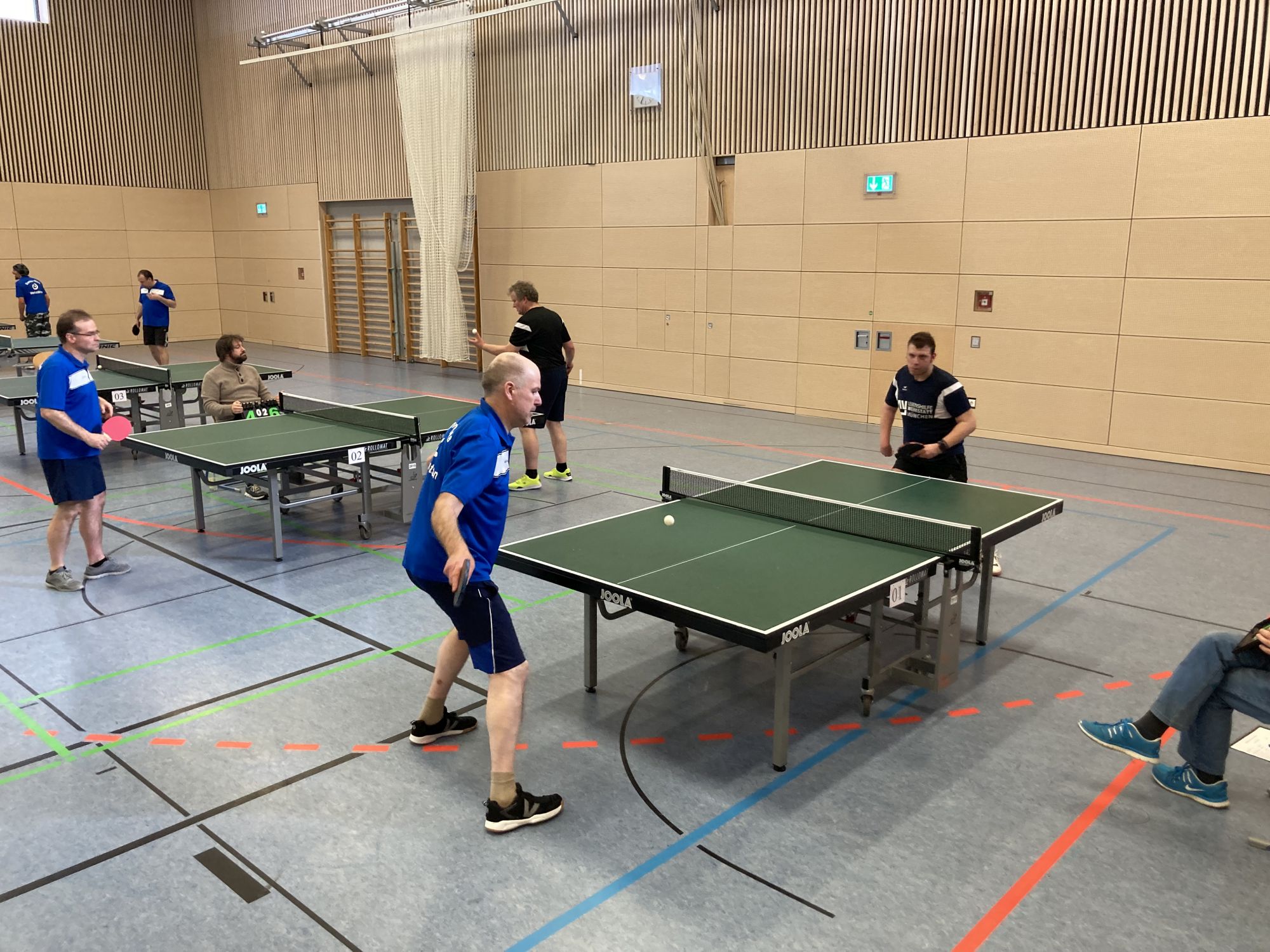 Tischtennisplatten in einem Raum an  welchen Männer mit blauem und schwarzem Dress gegeneinander  spielen