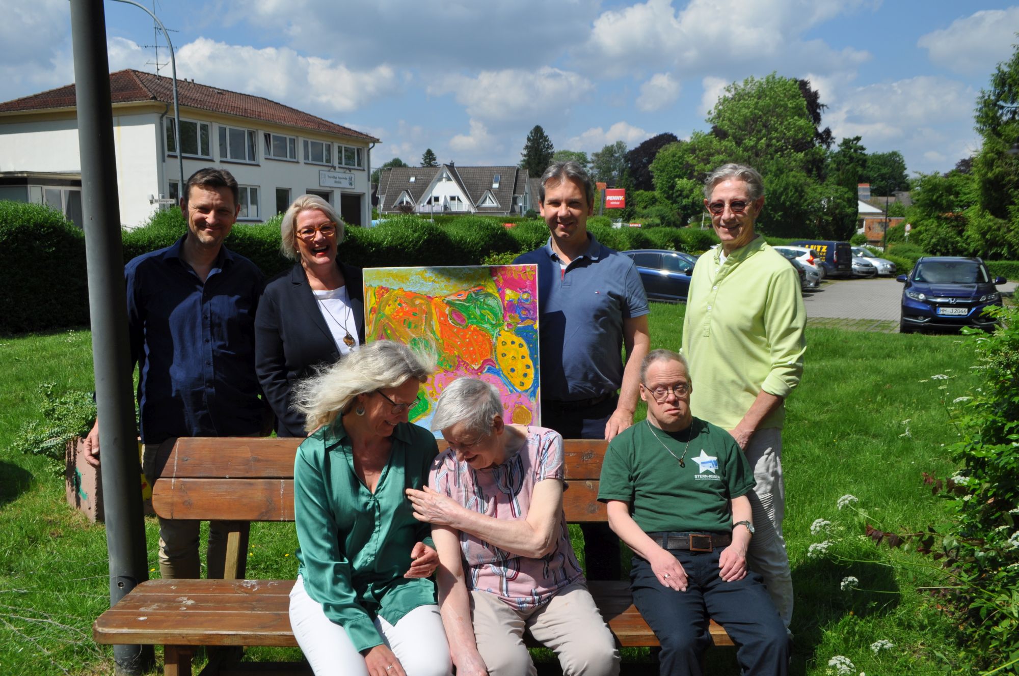 Im Hintergrunnd ein zweigeschossiges Gebäude davor  eine Bank mit drei Personen dahinter  vier Personen stehend