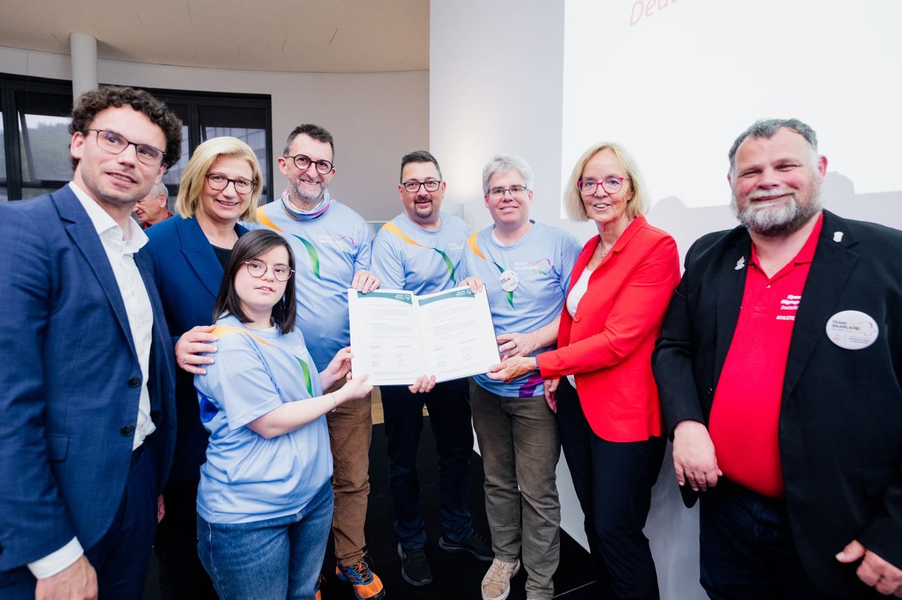 Gruppe von acht Männern  und Frauen steht in einer Zimmerecke und zweigt einen unterzeichneten Vertrag