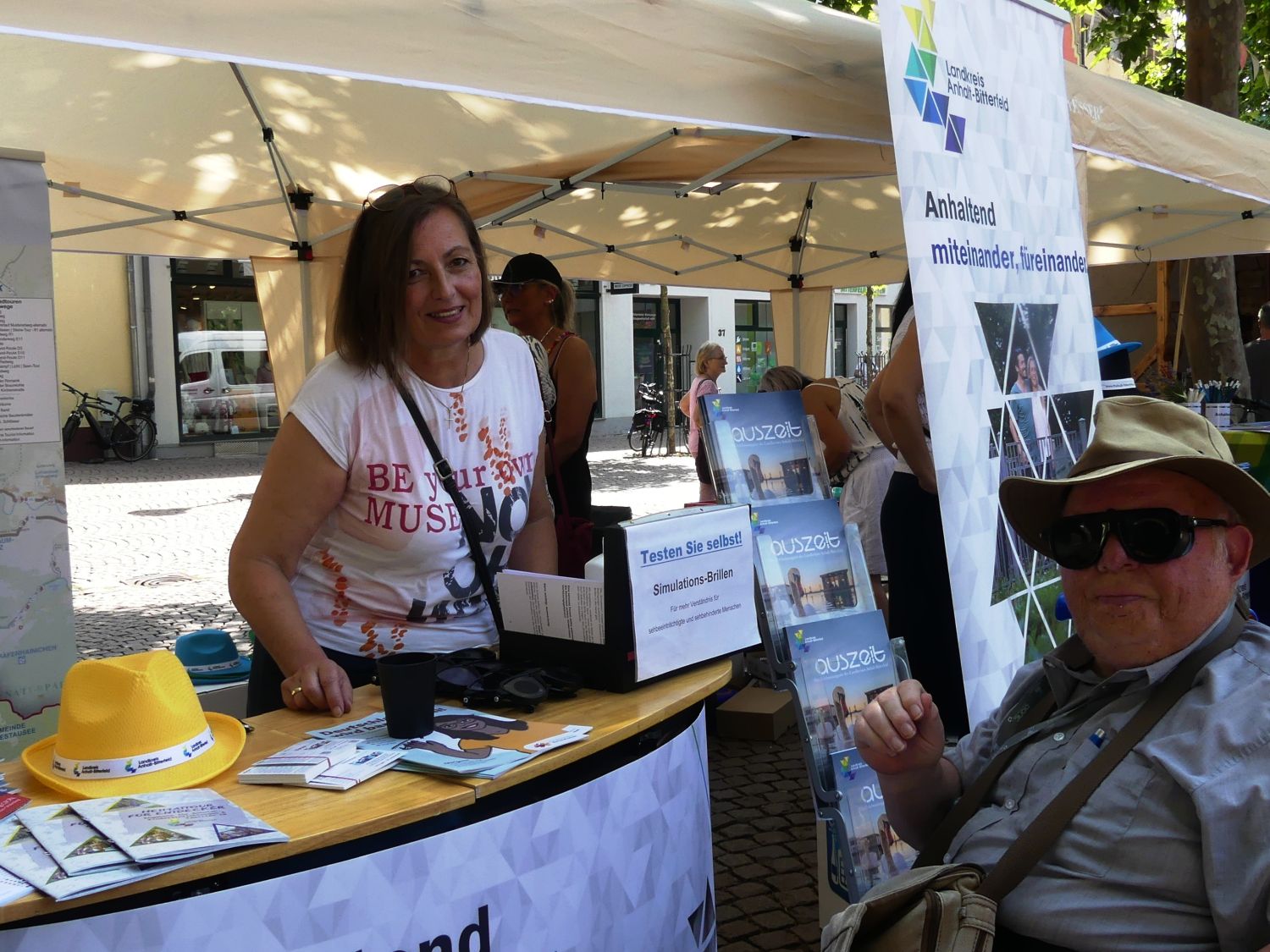 Junge Frau an  einem Stand mit vielen  Broschüren und einem Sortiment an Simulationsbrille im  Gespräch mit dem Autor