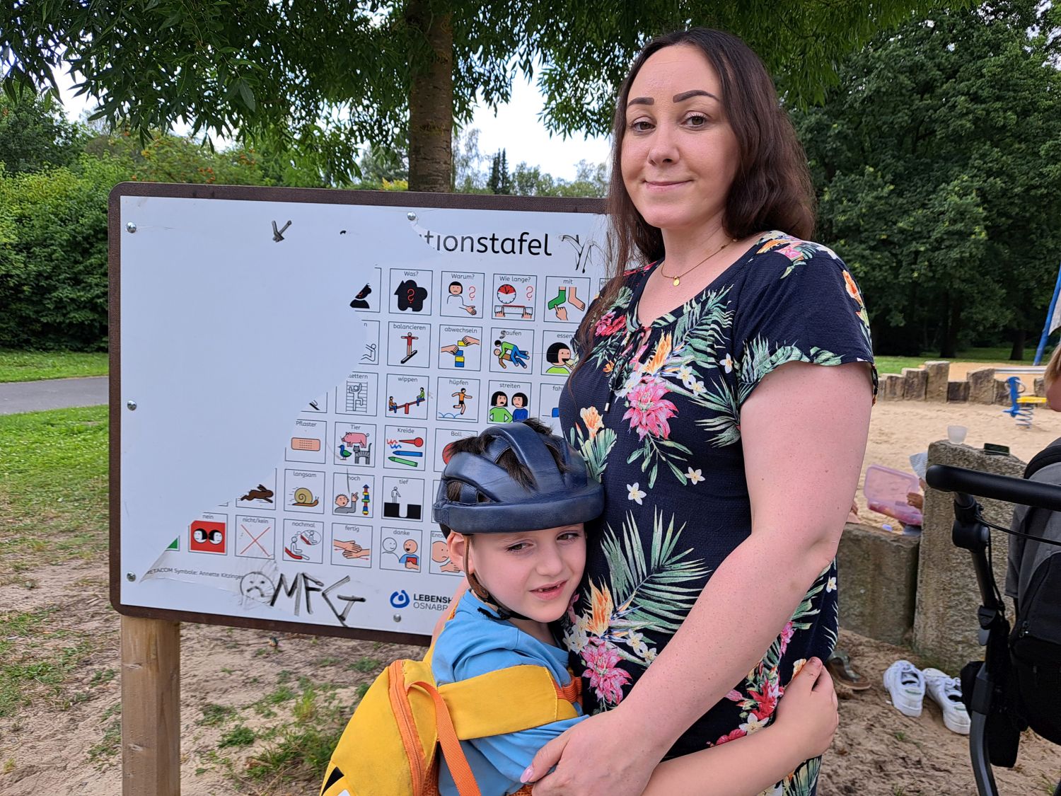 Kleiner Junge mit blauem Fahrradhelm und gelbem Rucksack nebem einer erwachsenen Frau vor der beschädigten Tafel