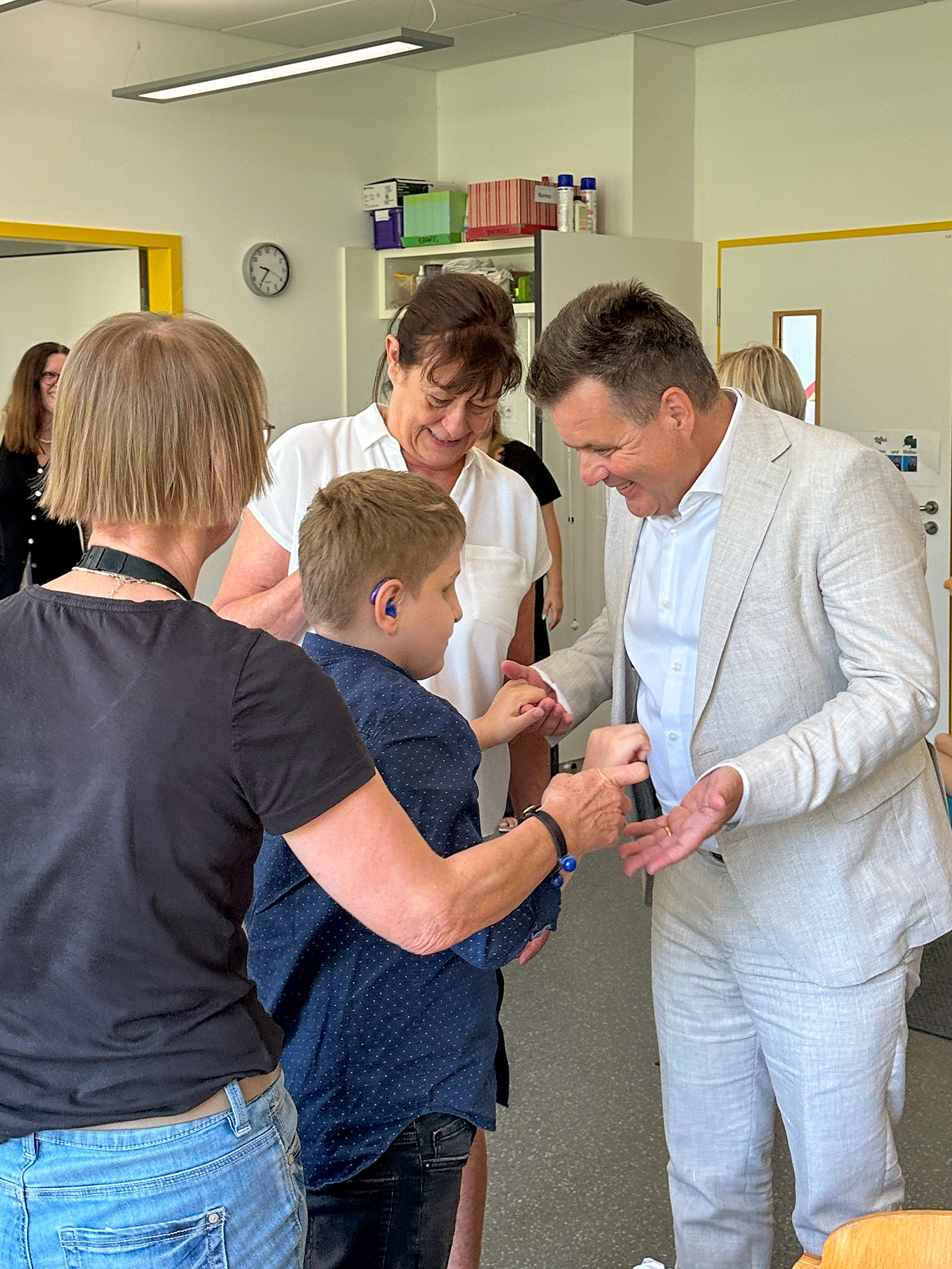 Ein Junge mit einem Hörgerät wird von einem Mann in einem hellen Anzug freundlich begrüß