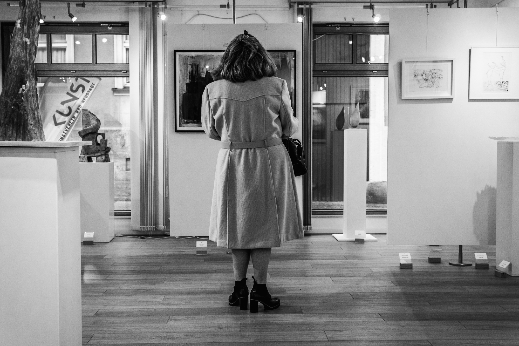Eine Frau steht mit dem Rücken zur Kamera in einem Ausstellungsraum und betrachtet Bilder und Skulpturen an den Wänden.