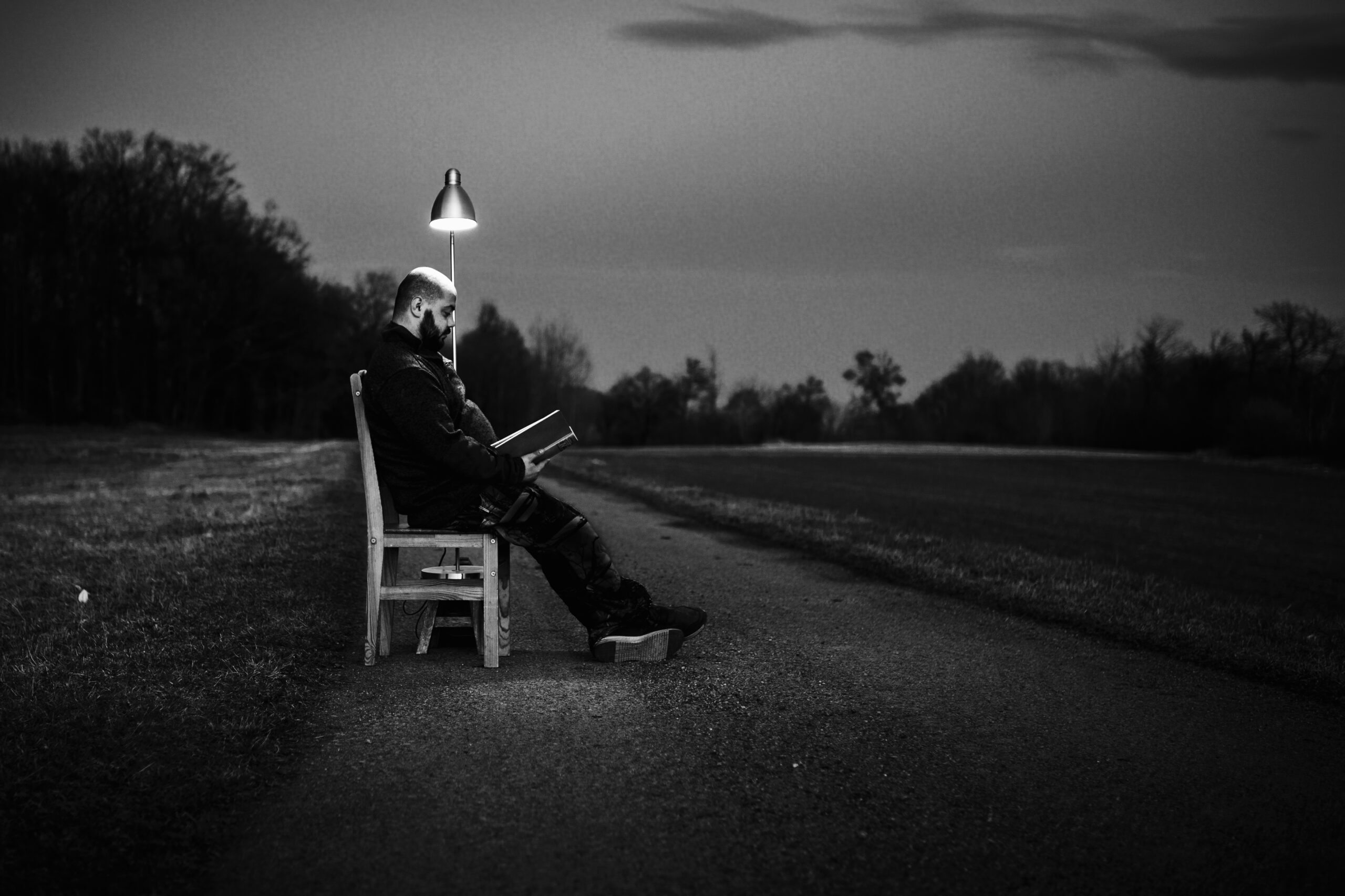 Schwarz-weißes Foto: Mann sitzt nachts allein auf einem Stuhl im Freien, liest in einem Buch, beleuchtet nur von einer einzelnen Schreibtischlampe – stille, surreale Szene.