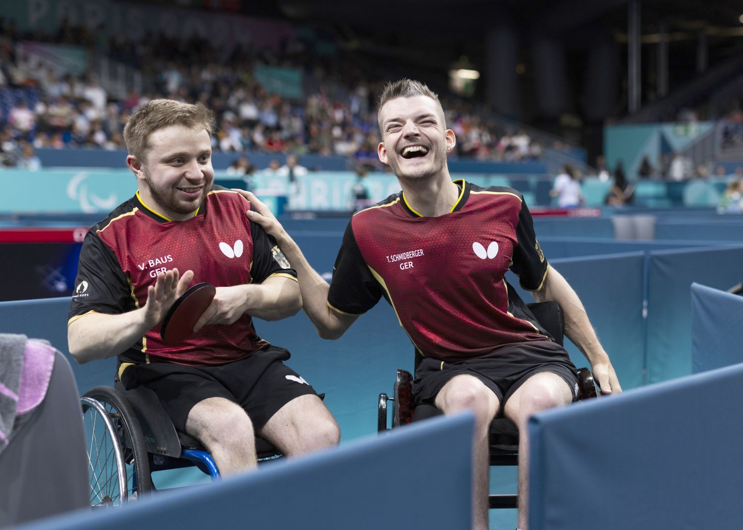 Zwei junge Tischtennisssspieler mit rot-schwarzem Trikot auf Rollstühlen an einer Tischtennisplatte mit Tischtennisschlägern in den Händen