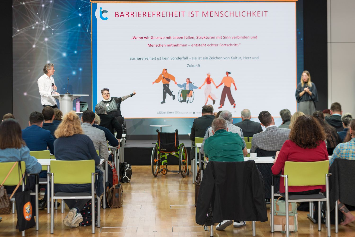 Blick auf eine Bühne mit drei Personen, davon eine mit Rollstuhl. Rückwand bildet Thema der Ko9nfernez Barrierefreiheit ist Menschlichkeit mit Text zu Tagung und Bild mit vier Personen, davon eine mit Rollstuhl, die sich an den Händen halten