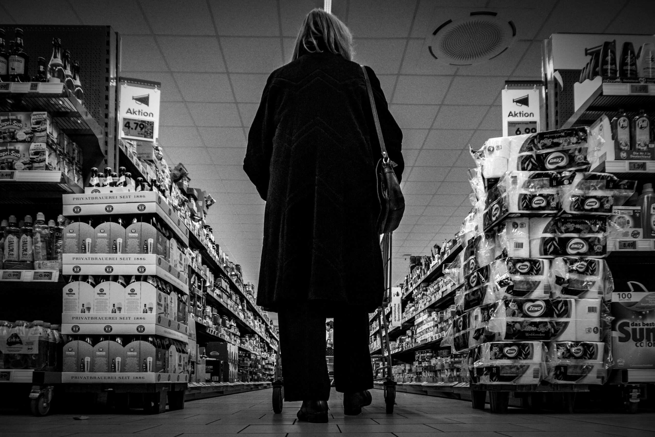Eine Frau von Hinten in einem Gang voller Warenregale in einem Lebensmittel-Discounter , schwarz-weiß Fotografie