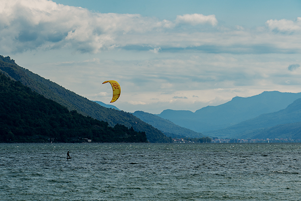 Wassersportler auf dem Wasser. Über ihm fliegt ein gelber Kite. Im Hintergrund befinden sich bewaldete Berge. Himmel ist leicht bewölkt.