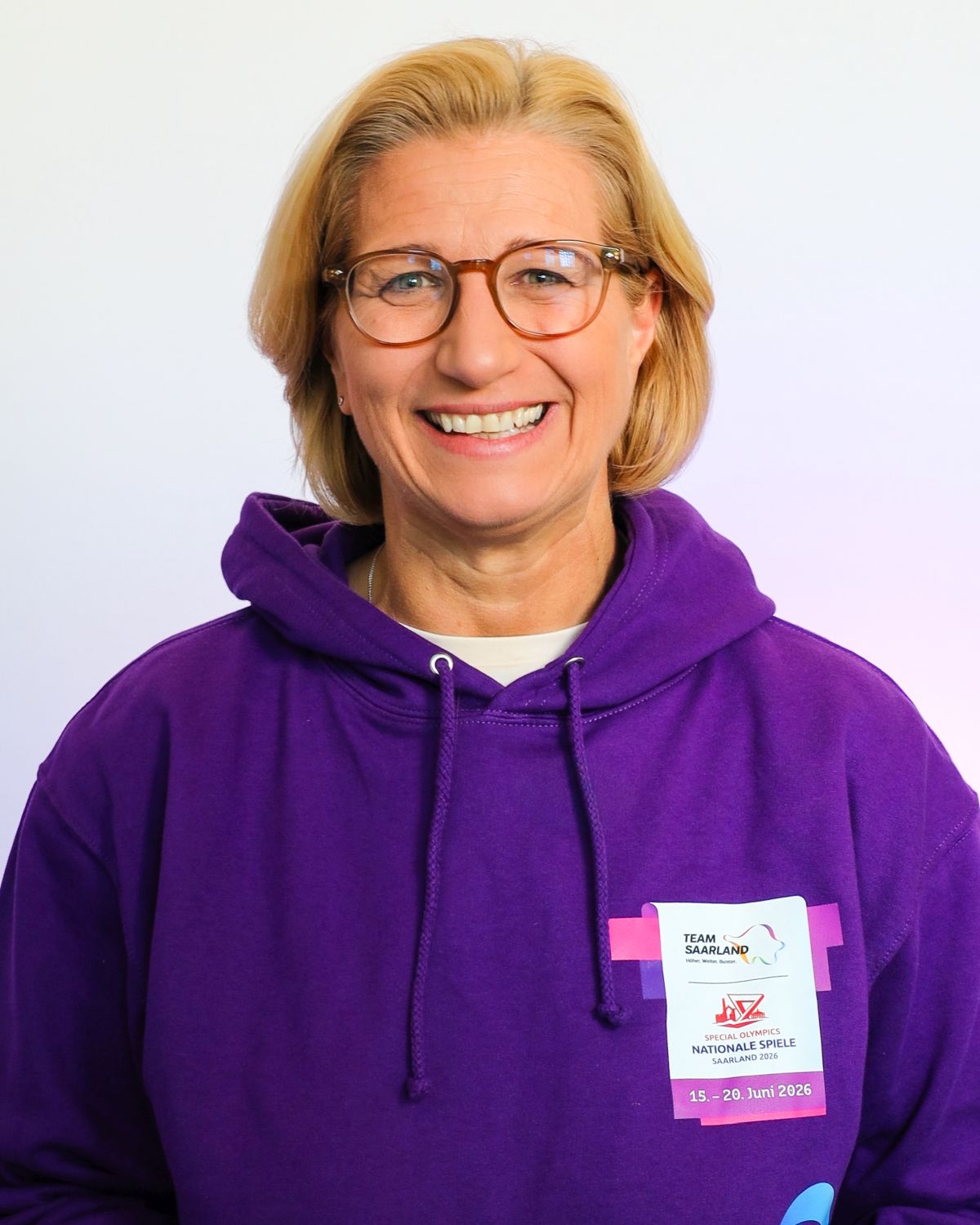 Brustbild einer Frau mit blonden schulterlangem Haar und Brille in lila Sportjacke