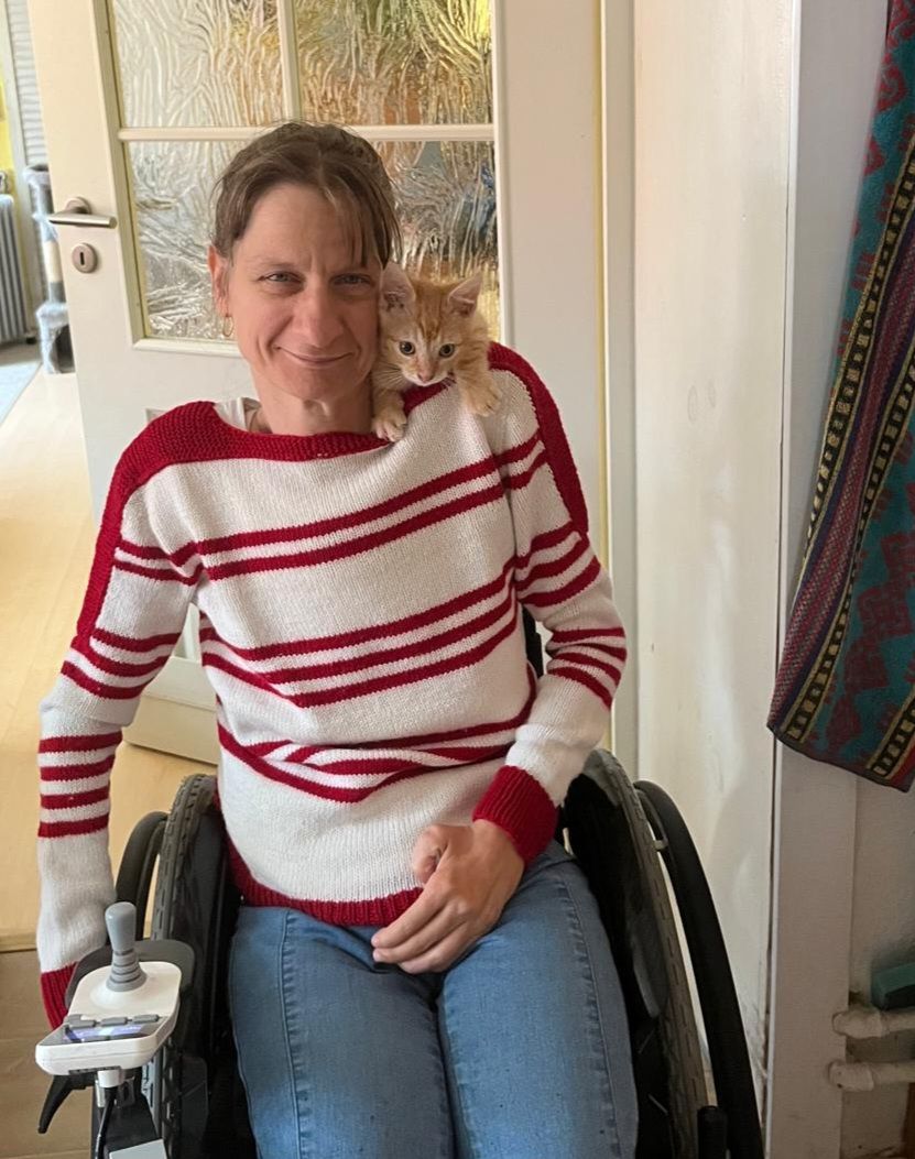 Junge Frau in weißem Pullover mit roten Querstreifen und Jeanshose in einem Rollstuhl. Auf der linken Schulter sitzt eine kleine Katze